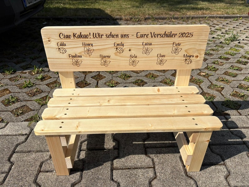 May include: A small, unfinished wooden bench with a backrest. The backrest has engraved text and names, along with butterfly and ladybug designs. The bench is constructed with visible screws and has a natural wood grain finish.