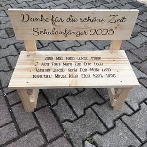 May include: A small, light-colored wooden bench with a backrest. The backrest has the text "Danke f&uuml;r die sch&ouml;ne Zeit Schulanf&auml;nger 2025" engraved on it. The bench seat has names engraved on it. A charming, personalized gift.