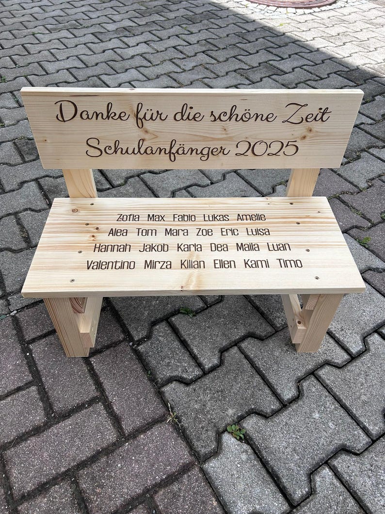 May include: A small, light-colored wooden bench with a backrest. The backrest has the text "Danke f&uuml;r die sch&ouml;ne Zeit Schulanf&auml;nger 2025" engraved on it. The bench seat has names engraved on it. A charming, personalized gift.