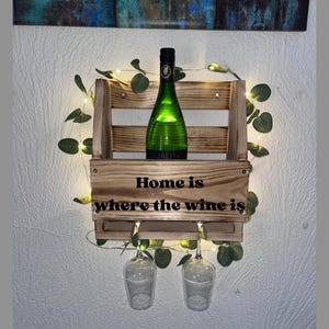 Puede incluir: Estante de vino de madera con una botella de vino verde y dos copas de vino. El estante tiene la frase "Home is where the wine is" y est&aacute; decorado con vegetaci&oacute;n y luces de hadas. Montado en una pared blanca.