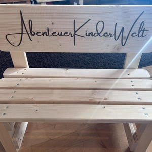 May include: A small, light-colored wooden bench with a backrest. The backrest features the cursive text "Abenteuer KinderWelt" in black. The bench has four horizontal slats for the seat and is constructed with visible screws.