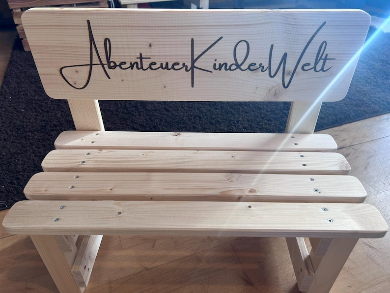 May include: A small, light-colored wooden bench with a backrest. The backrest features the cursive text "Abenteuer KinderWelt" in black. The bench has four horizontal slats for the seat and is constructed with visible screws.