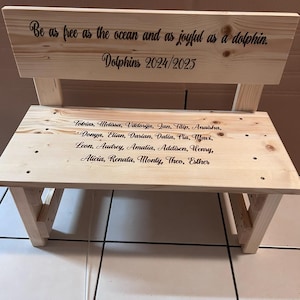 May include: A small, unfinished wooden bench with a backrest. The backrest has the text "Be as free as the ocean and as joyful as a dolphin. Dolphins 2024/2025." The bench seat has a list of names. The bench is made of light-colored wood.