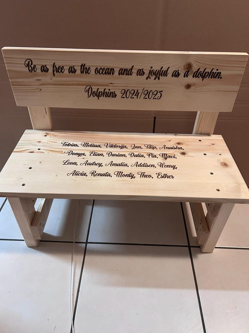 May include: A small, unfinished wooden bench with a backrest. The backrest has the text "Be as free as the ocean and as joyful as a dolphin. Dolphins 2024/2025." The bench seat has a list of names. The bench is made of light-colored wood.