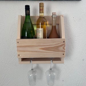 Puede incluir: Un botellero de madera clara, montado en la pared, con tres botellas y dos copas de vino. Las botellas son verdes, doradas y marrones. Las copas de vino son transparentes. El botellero est&aacute; fijado a una pared blanca.