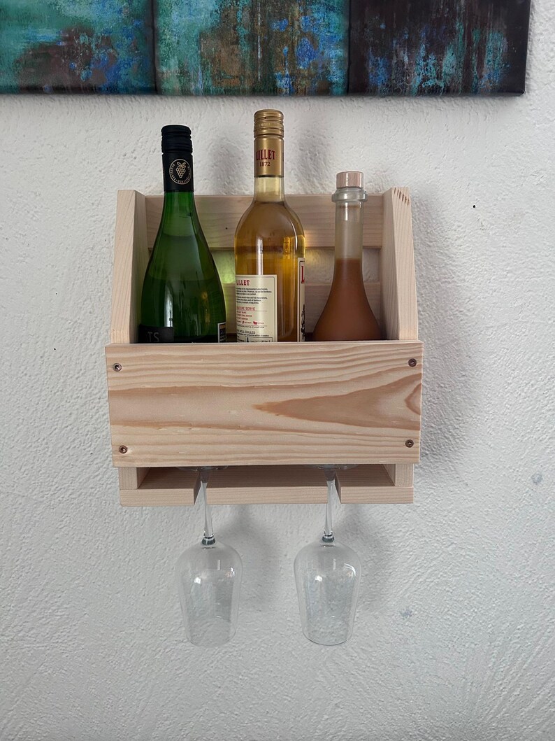 Puede incluir: Un botellero de madera clara, montado en la pared, con tres botellas y dos copas de vino. Las botellas son verdes, doradas y marrones. Las copas de vino son transparentes. El botellero est&aacute; fijado a una pared blanca.