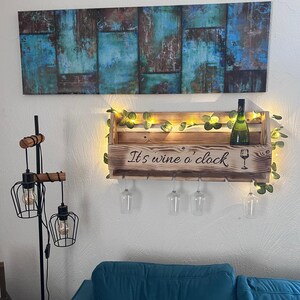 Puede incluir: Un botellero de madera con el texto "It's wine o'clock" y un gr&aacute;fico de copa de vino. El botellero contiene una botella de vino y cuatro copas de vino. El botellero est&aacute; decorado con luces de hadas y vegetaci&oacute;n. Encima cuelga un cuadro con tonos azules y marrones.