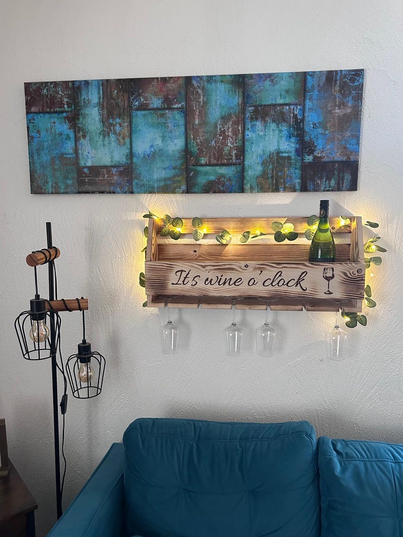 Puede incluir: Un botellero de madera con el texto "It's wine o'clock" y un gr&aacute;fico de copa de vino. El botellero contiene una botella de vino y cuatro copas de vino. El botellero est&aacute; decorado con luces de hadas y vegetaci&oacute;n. Encima cuelga un cuadro con tonos azules y marrones.