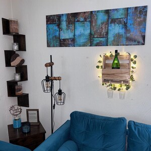 Puede incluir: Escena de sal&oacute;n con un sof&aacute; azul verdoso, una estanter&iacute;a esquinera de madera con velas y una pintura abstracta mural con tonos azules y marrones. Tambi&eacute;n se ve un botellero de madera con una botella verde y luces de hadas.