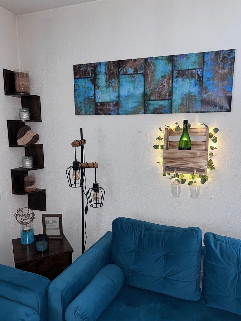 Puede incluir: Escena de sal&oacute;n con un sof&aacute; azul verdoso, una estanter&iacute;a esquinera de madera con velas y una pintura abstracta mural con tonos azules y marrones. Tambi&eacute;n se ve un botellero de madera con una botella verde y luces de hadas.