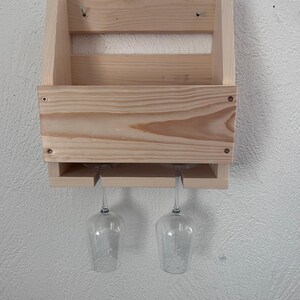 Puede incluir: Un botellero de madera natural, dise&ntilde;ado para guardar copas de vino. El botellero tiene un compartimento para botellas y una secci&oacute;n inferior con ranuras para dos copas. El botellero est&aacute; montado en una pared blanca.