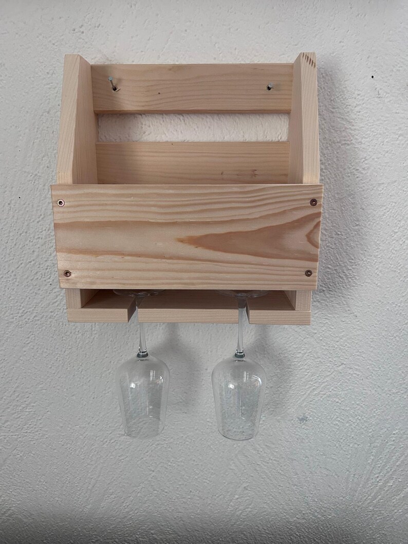 Puede incluir: Un botellero de madera natural, dise&ntilde;ado para guardar copas de vino. El botellero tiene un compartimento para botellas y una secci&oacute;n inferior con ranuras para dos copas. El botellero est&aacute; montado en una pared blanca.
