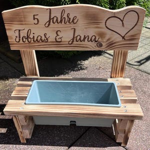K&ouml;nnte beinhalten: Holz-Pflanzbank mit hellblauem Beh&auml;lter. Die R&uuml;ckwand ist mit "5 Jahre Tobias & Jana" und einem Herz graviert. Die Bank ist aus hellem Holz gefertigt und f&uuml;r den Au&szlig;enbereich geeignet.