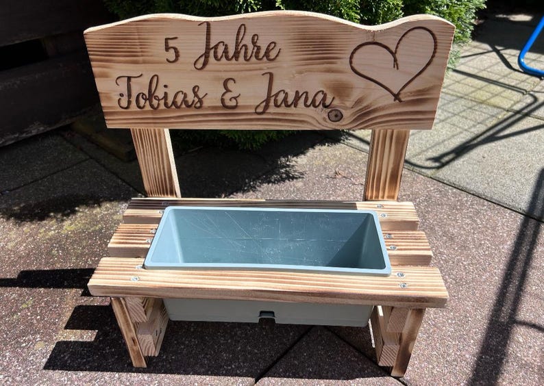 K&ouml;nnte beinhalten: Holz-Pflanzbank mit hellblauem Beh&auml;lter. Die R&uuml;ckwand ist mit "5 Jahre Tobias & Jana" und einem Herz graviert. Die Bank ist aus hellem Holz gefertigt und f&uuml;r den Au&szlig;enbereich geeignet.