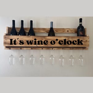 Puede incluir: Un botellero de madera con el texto "It's wine o'clock" en negro. El botellero contiene varias botellas de vino y copas de vino. El botellero es de madera marr&oacute;n claro y est&aacute; montado en una pared blanca.