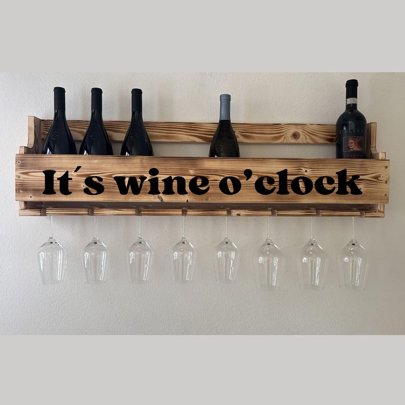 Puede incluir: Un botellero de madera con el texto "It's wine o'clock" en negro. El botellero contiene varias botellas de vino y copas de vino. El botellero es de madera marr&oacute;n claro y est&aacute; montado en una pared blanca.