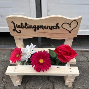 K&ouml;nnte beinhalten: Ein kleiner Holzbank-Pflanzk&uuml;bel mit dem Wort "Lieblingsmensch" und einem Herz-Ausschnitt. Der Pflanzk&uuml;bel enth&auml;lt rote, wei&szlig;e und rosa Kunstblumen. Die Bank ist aus hellem Holz gefertigt.