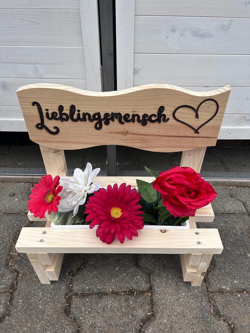 K&ouml;nnte beinhalten: Ein kleiner Holzbank-Pflanzk&uuml;bel mit dem Wort "Lieblingsmensch" und einem Herz-Ausschnitt. Der Pflanzk&uuml;bel enth&auml;lt rote, wei&szlig;e und rosa Kunstblumen. Die Bank ist aus hellem Holz gefertigt.