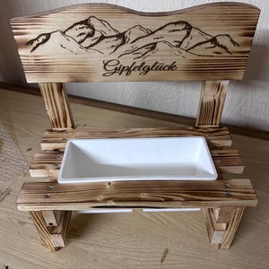 K&ouml;nnte beinhalten: Eine kleine Holzbank mit einem Bergketten-Design und dem Wort "Gipfelgl&uuml;ck" in die R&uuml;ckenlehne ge&auml;tzt. Sie verf&uuml;gt &uuml;ber einen wei&szlig;en rechteckigen Pflanzkasten. Die Bank ist aus hellbraunem Holz gefertigt.