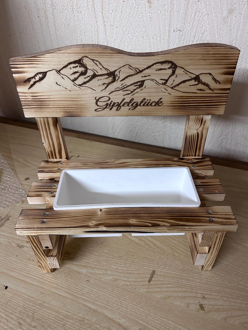 K&ouml;nnte beinhalten: Eine kleine Holzbank mit einem Bergketten-Design und dem Wort "Gipfelgl&uuml;ck" in die R&uuml;ckenlehne ge&auml;tzt. Sie verf&uuml;gt &uuml;ber einen wei&szlig;en rechteckigen Pflanzkasten. Die Bank ist aus hellbraunem Holz gefertigt.