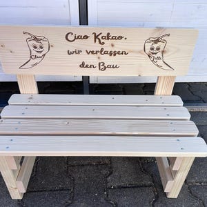May include: A small, light-colored wooden bench with a backrest. The backrest features the text "Ciao Kakao. wir verlassen den Bau" and two chili pepper illustrations. The bench has a slatted seat and angled legs, suitable for outdoor or indoor use.