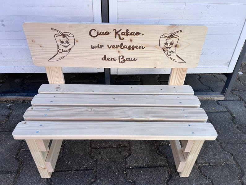 May include: A small, light-colored wooden bench with a backrest. The backrest features the text "Ciao Kakao. wir verlassen den Bau" and two chili pepper illustrations. The bench has a slatted seat and angled legs, suitable for outdoor or indoor use.