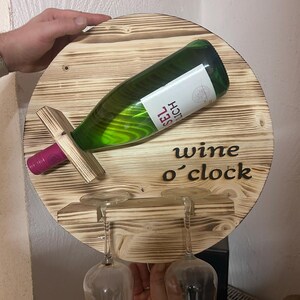 May include: A round wooden wall-mounted wine rack with the text "wine o'clock" burned into the wood. The rack holds a bottle of wine and two wine glasses.