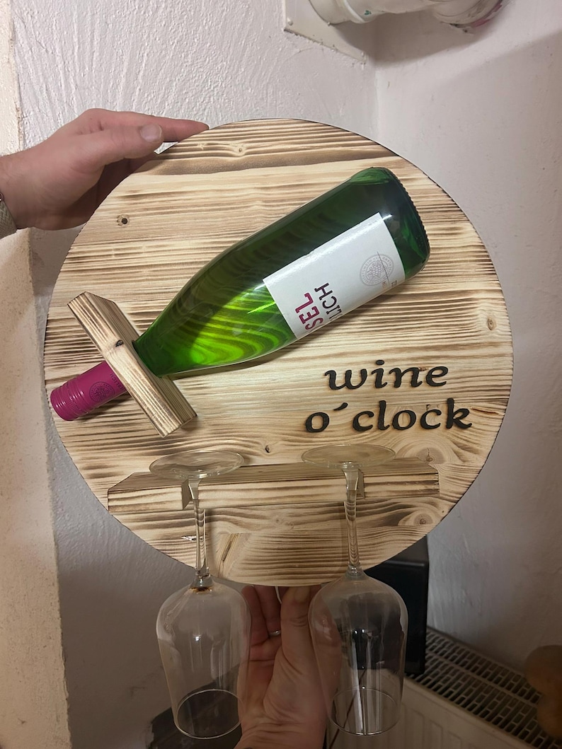 May include: A round wooden wall-mounted wine rack with the text "wine o'clock" burned into the wood. The rack holds a bottle of wine and two wine glasses.