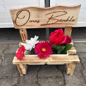 K&ouml;nnte beinhalten: Ein holzfarbener, bankf&ouml;rmiger Pflanzk&uuml;bel mit dem eingebrannten Schriftzug "Omas B&auml;nkle" auf der R&uuml;ckenlehne. Der Pflanzk&uuml;bel enth&auml;lt eine Vielzahl bunter Kunstblumen, darunter rote Gerbera, Rosen und wei&szlig;e Bl&uuml;ten. Die Bank ist aus hellem Holz.