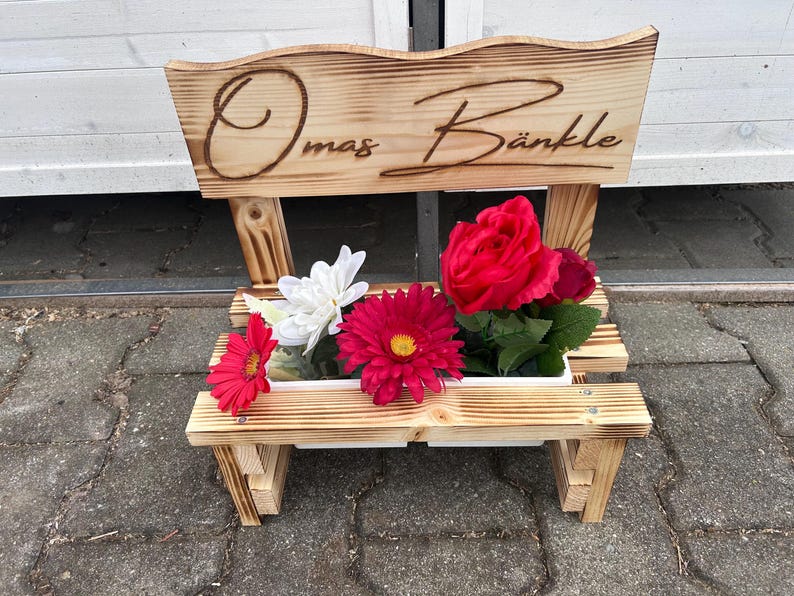 K&ouml;nnte beinhalten: Ein holzfarbener, bankf&ouml;rmiger Pflanzk&uuml;bel mit dem eingebrannten Schriftzug "Omas B&auml;nkle" auf der R&uuml;ckenlehne. Der Pflanzk&uuml;bel enth&auml;lt eine Vielzahl bunter Kunstblumen, darunter rote Gerbera, Rosen und wei&szlig;e Bl&uuml;ten. Die Bank ist aus hellem Holz.