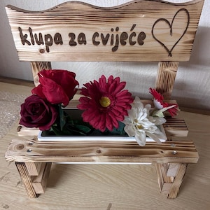 K&ouml;nnte beinhalten: Holzbank-Pflanzgef&auml;&szlig; mit dem Text "klupa za cvijeće" und einem Herzdesign. Die Bank enth&auml;lt rote und wei&szlig;e Kunstblumen. Die Bank ist aus hellbraunem Holz gefertigt.