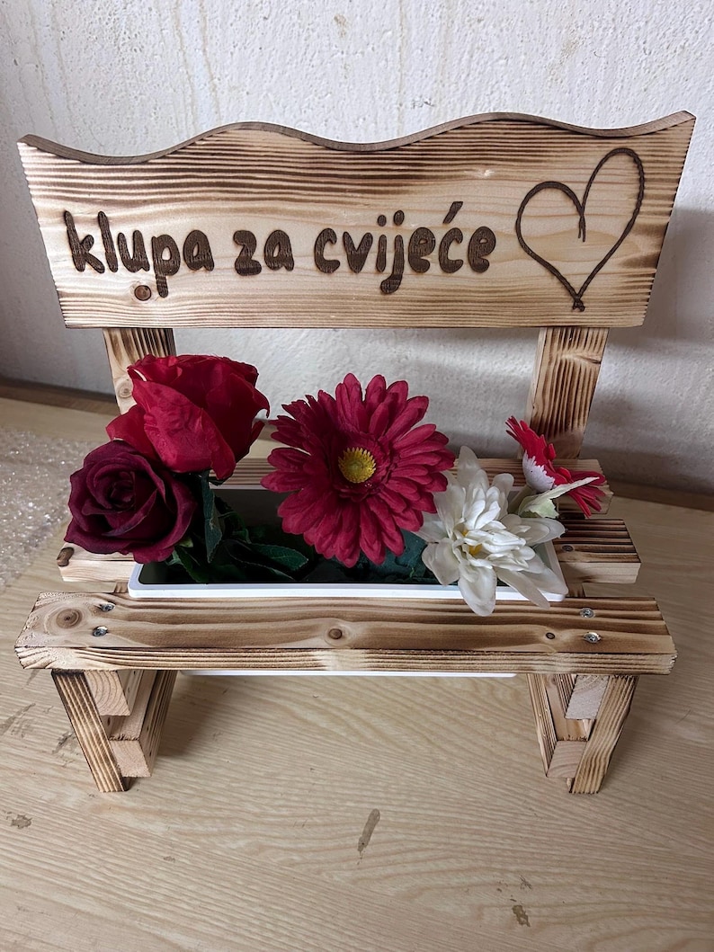 K&ouml;nnte beinhalten: Holzbank-Pflanzgef&auml;&szlig; mit dem Text "klupa za cvijeće" und einem Herzdesign. Die Bank enth&auml;lt rote und wei&szlig;e Kunstblumen. Die Bank ist aus hellbraunem Holz gefertigt.