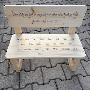 May include: A small, light-colored wooden bench with a backrest. The backrest has engraved text, and the seat has names engraved. The bench is made of unfinished wood and has a simple, classic design. The bench is outdoors.