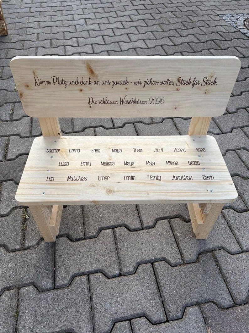 May include: A small, light-colored wooden bench with a backrest. The backrest has engraved text, and the seat has names engraved. The bench is made of unfinished wood and has a simple, classic design. The bench is outdoors.
