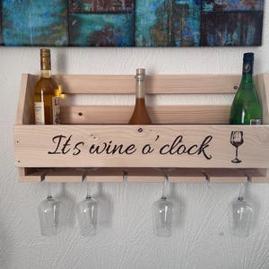 Puede incluir: Estante de vino de madera con el texto "It's wine o' clock" y un gr&aacute;fico de copa de vino. El estante tiene capacidad para tres botellas y cuatro copas de vino. Las botellas son de varios colores y formas. El estante est&aacute; montado en una pared blanca.