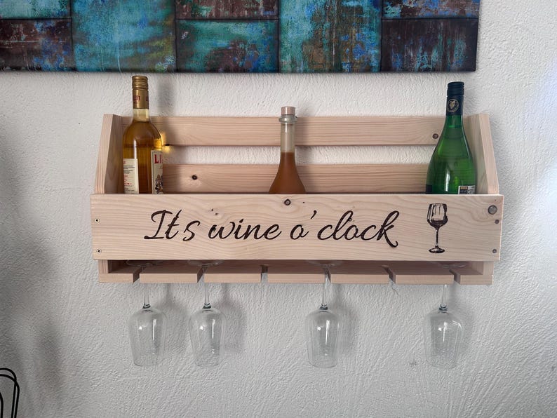 Puede incluir: Estante de vino de madera con el texto "It's wine o' clock" y un gr&aacute;fico de copa de vino. El estante tiene capacidad para tres botellas y cuatro copas de vino. Las botellas son de varios colores y formas. El estante est&aacute; montado en una pared blanca.