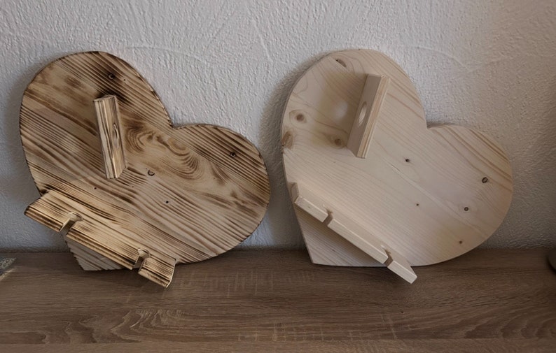 May include: Two wooden heart-shaped shelves with a rustic finish. One shelf has a natural wood color and the other has a darker, burnt wood finish. Both shelves have a small, rectangular shelf attached to the side.