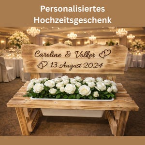Op de afbeelding: Een houten bank met een plantenbak gevuld met witte rozen. De bank heeft een rugleuning met de gegraveerde tekst "Caroline & Volker" en "13 augustus 2024". De bank is een gepersonaliseerd huwelijkscadeau.