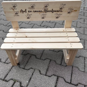 May include: A small, unfinished wooden bench with a backrest. The backrest features engraved bees and names, along with the phrase "Auf zu neuen Abenteuern." The bench has a slatted seat and is constructed with visible screws.