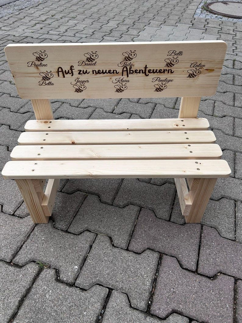 May include: A small, unfinished wooden bench with a backrest. The backrest features engraved bees and names, along with the phrase "Auf zu neuen Abenteuern." The bench has a slatted seat and is constructed with visible screws.