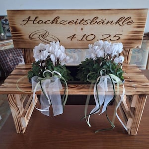 Może przedstawiać: Drewniana ławka z napisem "Hochzeitsb&auml;nkle 4.10.25" jest ozdobiona białymi kwiatami i zielonymi liśćmi. Ławka ma rustykalny design i jest ozdobiona białymi i zielonymi wstążkami.