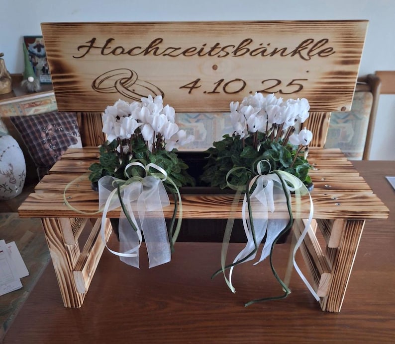 Może przedstawiać: Drewniana ławka z napisem "Hochzeitsb&auml;nkle 4.10.25" jest ozdobiona białymi kwiatami i zielonymi liśćmi. Ławka ma rustykalny design i jest ozdobiona białymi i zielonymi wstążkami.