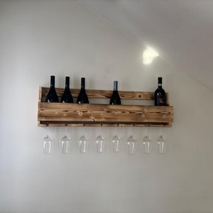 Puede incluir: Un botellero de madera r&uacute;stica con tinte oscuro, que contiene varias botellas de vino y copas. El botellero est&aacute; montado en una pared blanca, mostrando un dise&ntilde;o sencillo y elegante para el almacenamiento y la exhibici&oacute;n del vino.