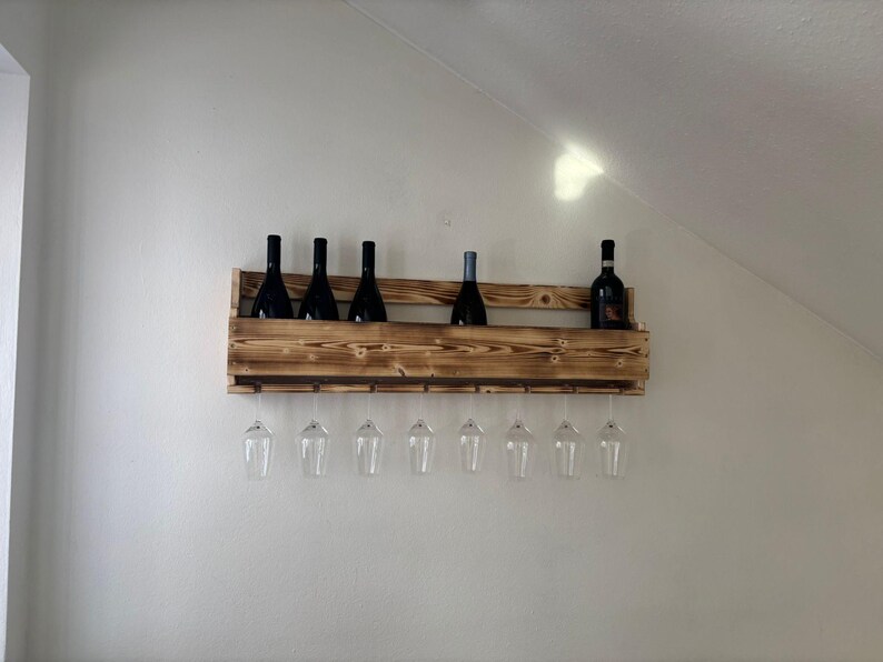 Puede incluir: Un botellero de madera r&uacute;stica con tinte oscuro, que contiene varias botellas de vino y copas. El botellero est&aacute; montado en una pared blanca, mostrando un dise&ntilde;o sencillo y elegante para el almacenamiento y la exhibici&oacute;n del vino.