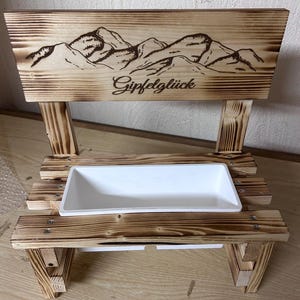 K&ouml;nnte beinhalten: Ein Holzpflanzer mit einem Bergketten-Design und dem Wort "Gipfelgl&uuml;ck" auf der R&uuml;ckseite. Der Pflanzer hat einen wei&szlig;en rechteckigen Beh&auml;lter f&uuml;r Pflanzen. Das Holz hat eine gebrannte Oberfl&auml;che.
