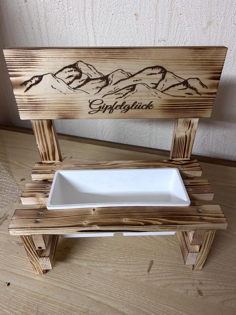 K&ouml;nnte beinhalten: Ein Holzpflanzer mit einem Bergketten-Design und dem Wort "Gipfelgl&uuml;ck" auf der R&uuml;ckseite. Der Pflanzer hat einen wei&szlig;en rechteckigen Beh&auml;lter f&uuml;r Pflanzen. Das Holz hat eine gebrannte Oberfl&auml;che.