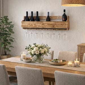 Puede incluir: Un comedor con una mesa de madera preparada para una comida. Una estanter&iacute;a de vino de madera contiene botellas y copas de vino. Un jarr&oacute;n con flores blancas est&aacute; sobre la mesa, junto con velas y objetos decorativos. Una planta grande est&aacute; en el fondo.