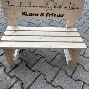 May include: A small, unfinished wooden bench with a backrest. The backrest has engraved text that reads "F&uuml;r viele sch&ouml;ne erste Schritte im Leben" and the names "Klara & Frieda". The bench is made of light-colored wood.