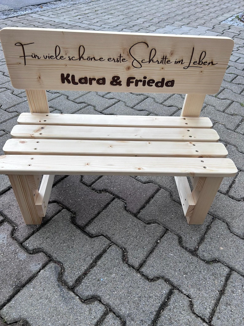 May include: A small, unfinished wooden bench with a backrest. The backrest has engraved text that reads "F&uuml;r viele sch&ouml;ne erste Schritte im Leben" and the names "Klara & Frieda". The bench is made of light-colored wood.