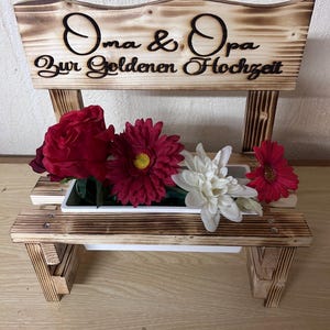 K&ouml;nnte beinhalten: Ein holzbankf&ouml;rmiger Pflanzk&uuml;bel mit den Worten "Oma & Opa Zur Goldenen Hochzeit" in schwarzer Schrift. Der Pflanzk&uuml;bel enth&auml;lt rote, wei&szlig;e und rosa Blumen. Das Holz hat eine gebrannte Oberfl&auml;che.